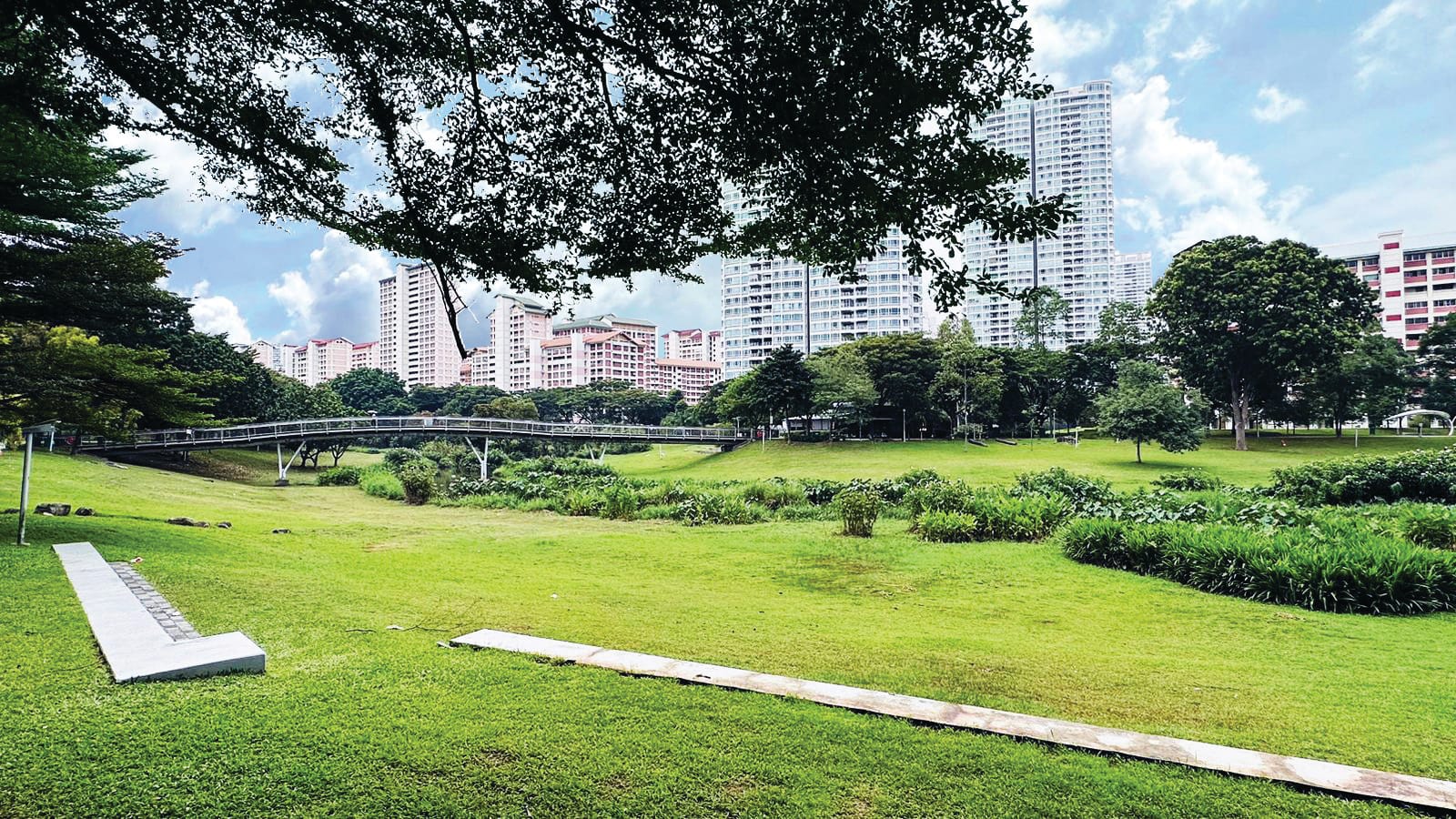 Park with green lawn and trees, a bridge crossing a dip, and apartment buildings in the background under a cloudy sky.
